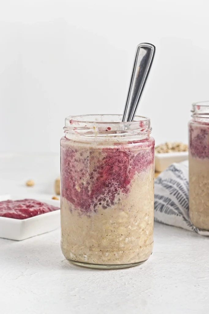 A glass jar filled with peanut butter and jam overnight oats, swirled with red fruit puree and topped with a spoon, sits on a white surface.