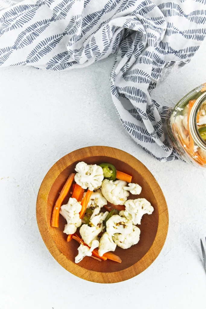 A wooden bowl with pickled cauliflower and carrots, jalapeños, on a white surface next to a patterned cloth and a glass jar.
