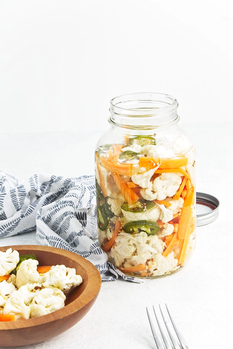A glass jar filled with pickled cauliflower and carrots, plus jalapeños, sits beside a wooden bowl with a serving and two forks on a light surface.