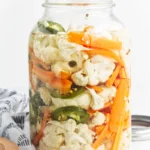 A glass jar filled with pickled cauliflower and carrots, along with sliced jalapeños, sits on a white surface beside a patterned cloth.