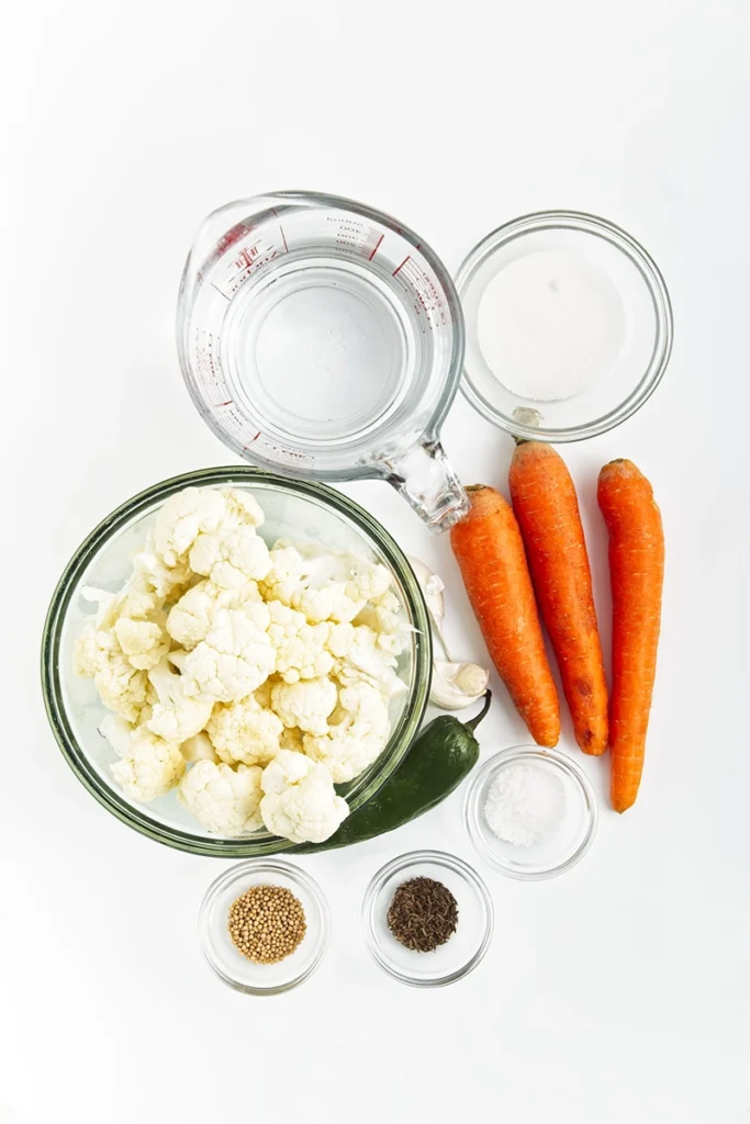 Overhead view of raw pickled cauliflower and carrots, a jalapeño, garlic cloves, water, sugar, salt, mustard seeds, and black peppercorns arranged in glass bowls on a white background.