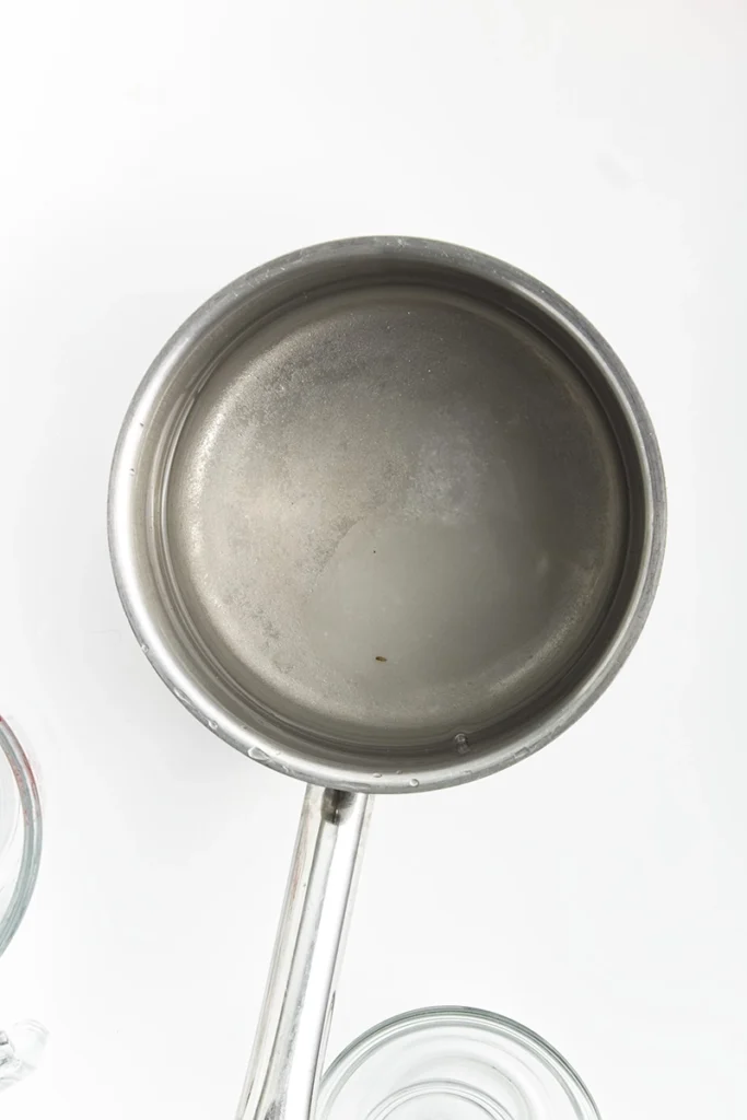 Overhead view of an empty stainless steel saucepan on a white surface, with part of a glass bowl of pickled cauliflower and carrots visible beside it.