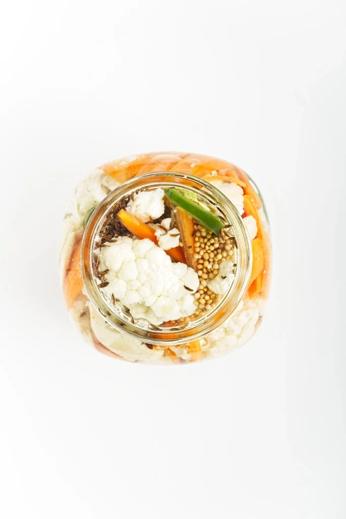 A top-down view of a glass jar filled with pickled cauliflower and carrots, green chili, and mustard seeds on a white background.