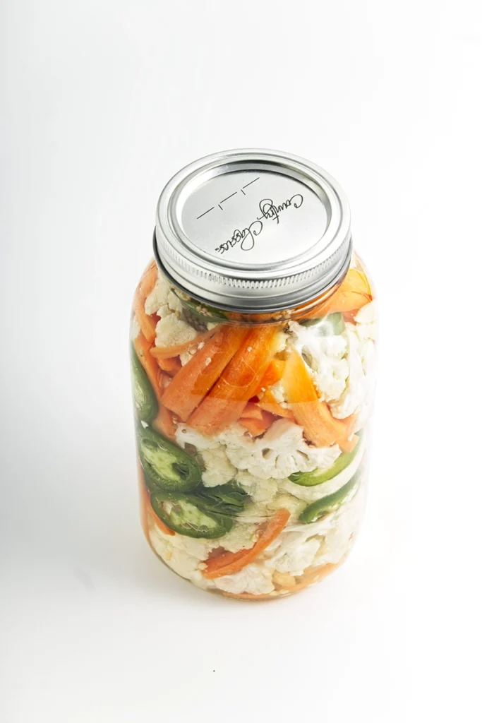 A glass jar filled with pickled cauliflower and carrots, jalapeños, and sealed with a metal lid sits on a white background.