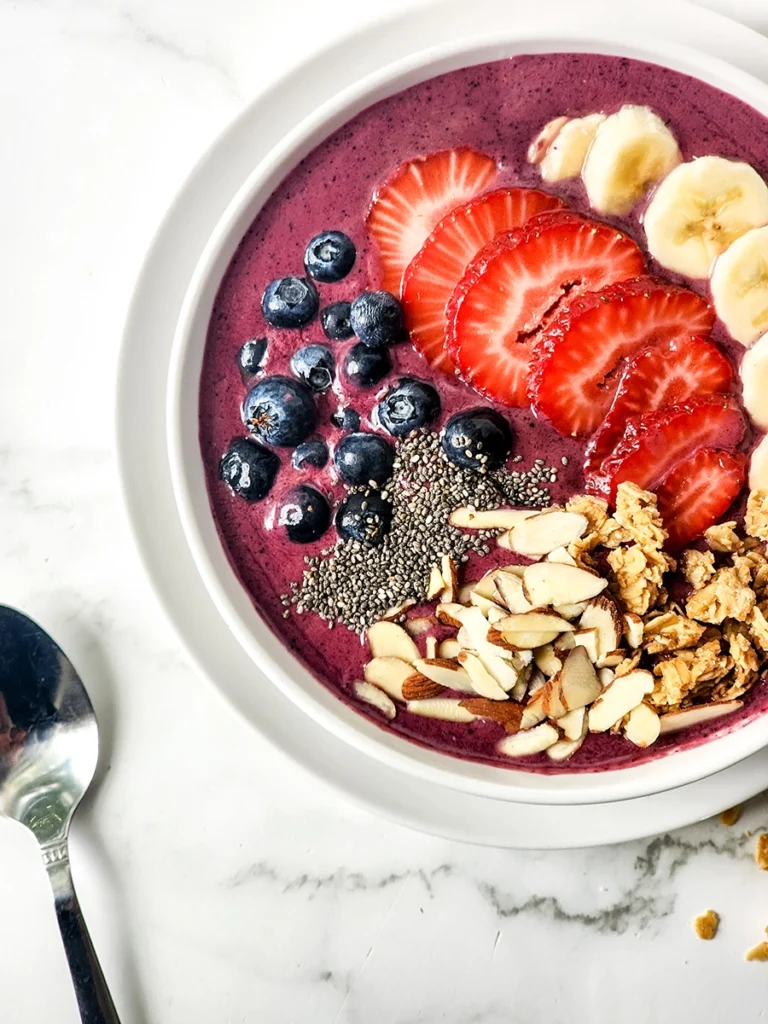 A single serving acai bowl topped with strawberries, bananas, blueberries, granola, chia seeds, and sliced almonds sits on a white plate beside a spoon on a marble surface.