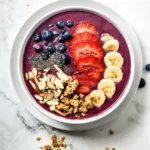 A single serving acai bowl topped with blueberries, sliced strawberries, banana, chia seeds, granola, and almond slices on a white marble surface.