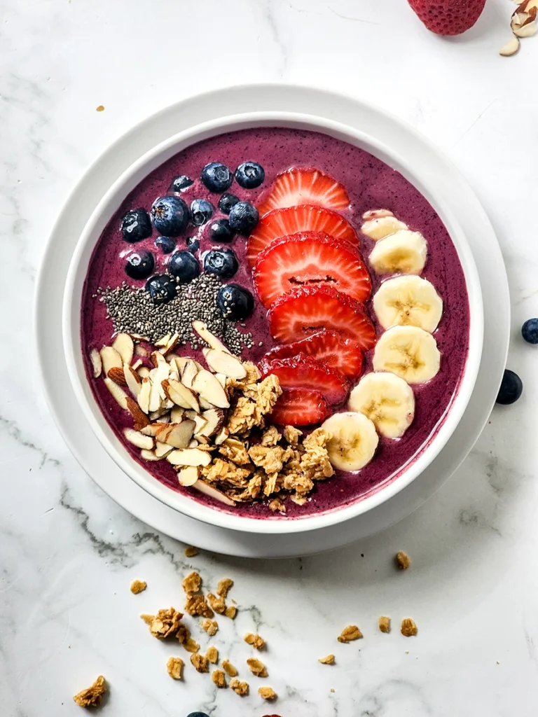 A single serving acai bowl topped with blueberries, sliced strawberries, banana, chia seeds, granola, and almond slices on a white marble surface.