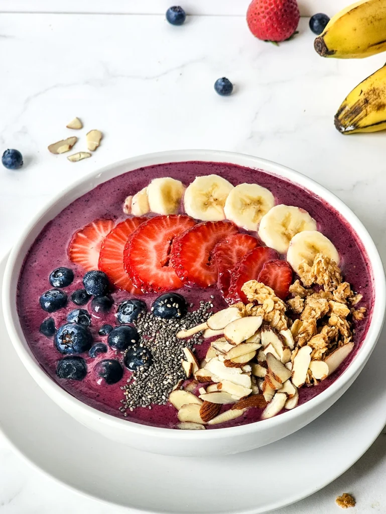 A single serving acai bowl topped with sliced bananas, strawberries, blueberries, granola, chia seeds, and almond slices on a white plate.