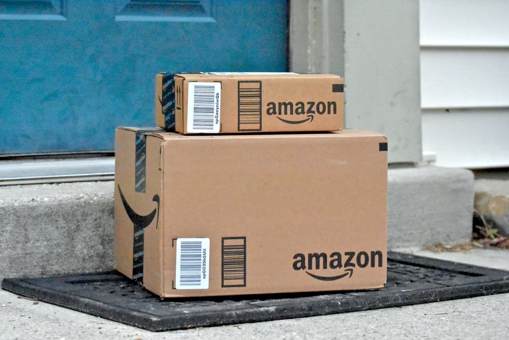Two Amazon packages—filled with things I actually use—are stacked on a doormat in front of a blue door and white siding.