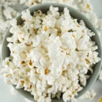 A bowl filled with plain microwave popcorn sits on a white surface with scattered popcorn pieces around it, making for a simple, single serving snack.