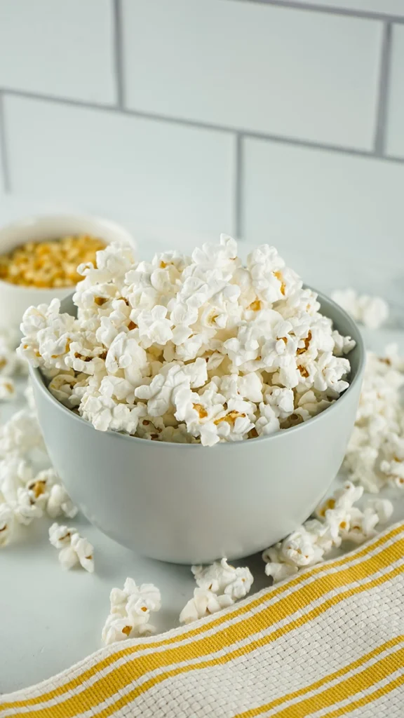 A white bowl filled with popped snack popcorn sits on a white surface, with some popcorn scattered around and a bowl of unpopped kernels in the background, perfect for single serve enjoyment.