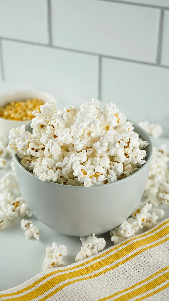 A white bowl filled with popped snack popcorn sits on a white surface, with some popcorn scattered around and a bowl of unpopped kernels in the background, perfect for single serve enjoyment.