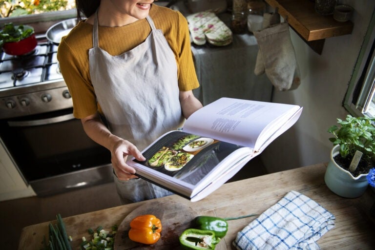 Person wearing an apron stands in a kitchen, holding open a cooking for one cookbook with food photos. Vegetables and a dish towel are on the wooden counter.