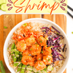 A bowl of rice topped with shrimp in orange sauce, sliced cucumbers, snap peas, shredded carrots, and shredded purple cabbage on a wooden surface creates the ultimate bang bang shrimp bowl. Text reads "Bang Bang Shrimp." If you’re looking for an easy shrimp bowl recipe packed with flavor and color, this is the perfect choice.