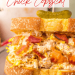 Enjoy a delicious chicken salad sandwich inspired by the famous Chicken Salad Chick Sassy Scotty recipe, featuring savory chicken salad with bacon and shredded cheese on thick bread, topped with a pickle and beautifully displayed on a plate.