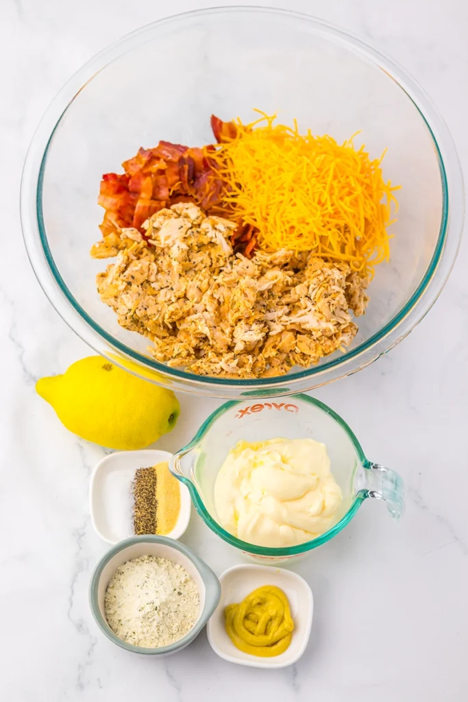 A glass bowl with shredded chicken, bacon, and shredded cheese—perfect for recreating the famous Chicken Salad Chick Sassy Scotty recipe—sits next to a lemon, mayonnaise, mustard, ranch seasoning, and black pepper on a white surface.