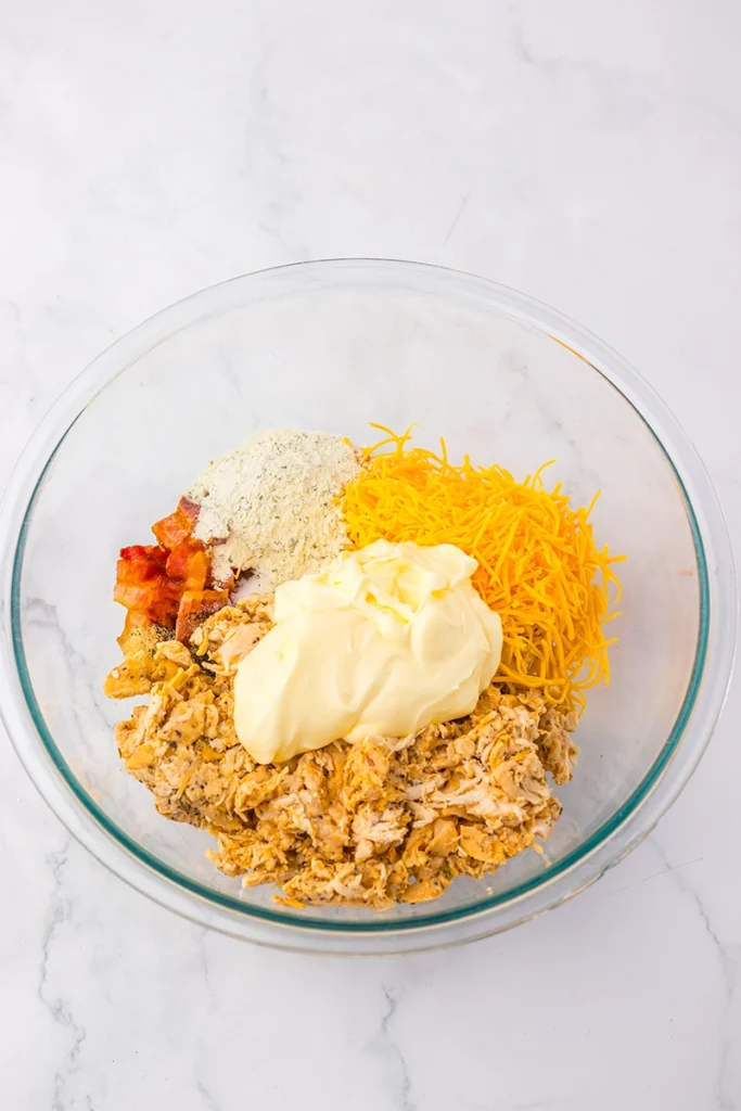 A glass bowl with shredded chicken, shredded cheddar cheese, mayonnaise, bacon bits, and ranch seasoning on a white marble surface—perfect for recreating the famous Chicken Salad Chick Sassy Scotty recipe at home.