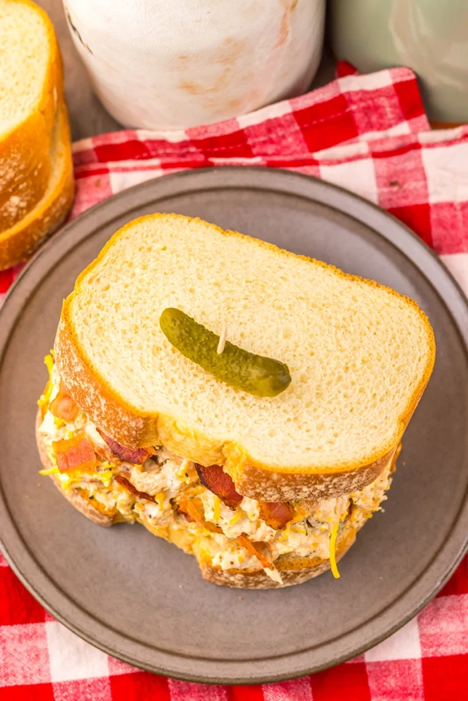 Chicken Salad Chick Sassy Scotty chicken salad sandwich with bacon and shredded cheese on white bread, topped with a pickle slice and secured with a toothpick, served on a gray plate over a red and white checkered cloth. OR Inspired by the Chicken Salad Chick Sassy Scotty recipe, this chicken salad sandwich features bacon and shredded cheese on white bread, topped with a pickle slice and secured with.