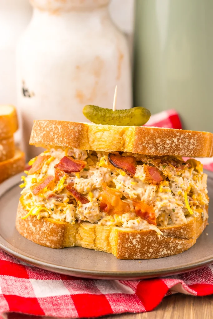 Chicken Salad Chick Sassy Scotty chicken salad sandwich with bacon and shredded cheese on sliced bread, topped with a small pickle on a toothpick, served on a gray plate with a red and white checkered napkin underneath.