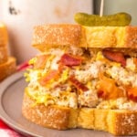 A sandwich featuring Chicken Salad Chick Sassy Scotty chicken salad, crisp bacon, and melted cheese on thick bread, topped with a pickle slice on a toothpick, served on a gray plate with a red and white napkin underneath.