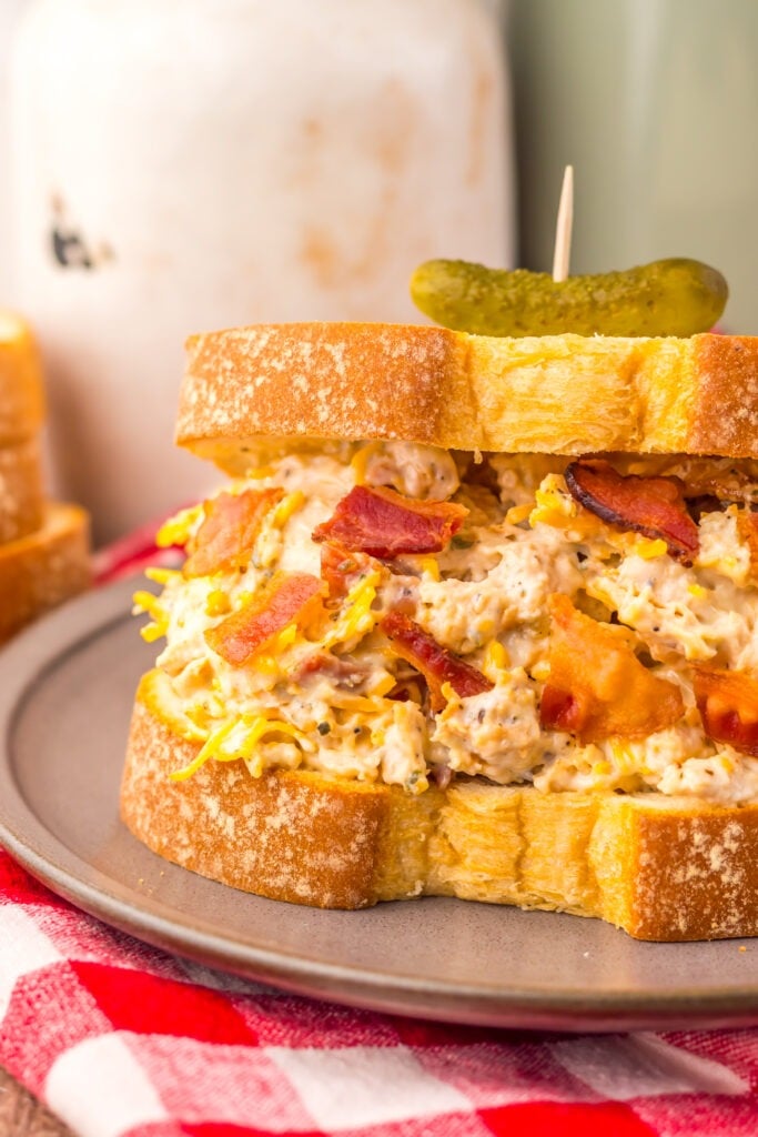 A sandwich featuring Chicken Salad Chick Sassy Scotty chicken salad, crisp bacon, and melted cheese on thick bread, topped with a pickle slice on a toothpick, served on a gray plate with a red and white napkin underneath.