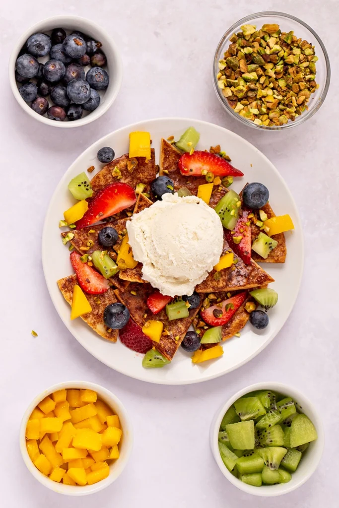 A delightful plate of fruit nachos for 1, topped with creamy vanilla ice cream, surrounded by bowls of blueberries, chopped pistachios, diced mango, and sliced kiwi.
