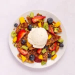A white plate with fruit nachos for 1, featuring chips topped with strawberries, blueberries, kiwi, and mango, garnished with pistachios and a scoop of vanilla ice cream in the center.