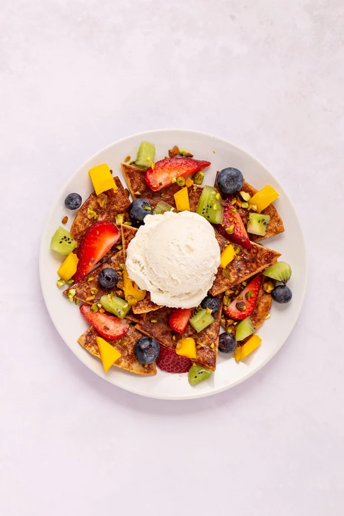 A white plate with fruit nachos for 1, featuring chips topped with strawberries, blueberries, kiwi, and mango, garnished with pistachios and a scoop of vanilla ice cream in the center.