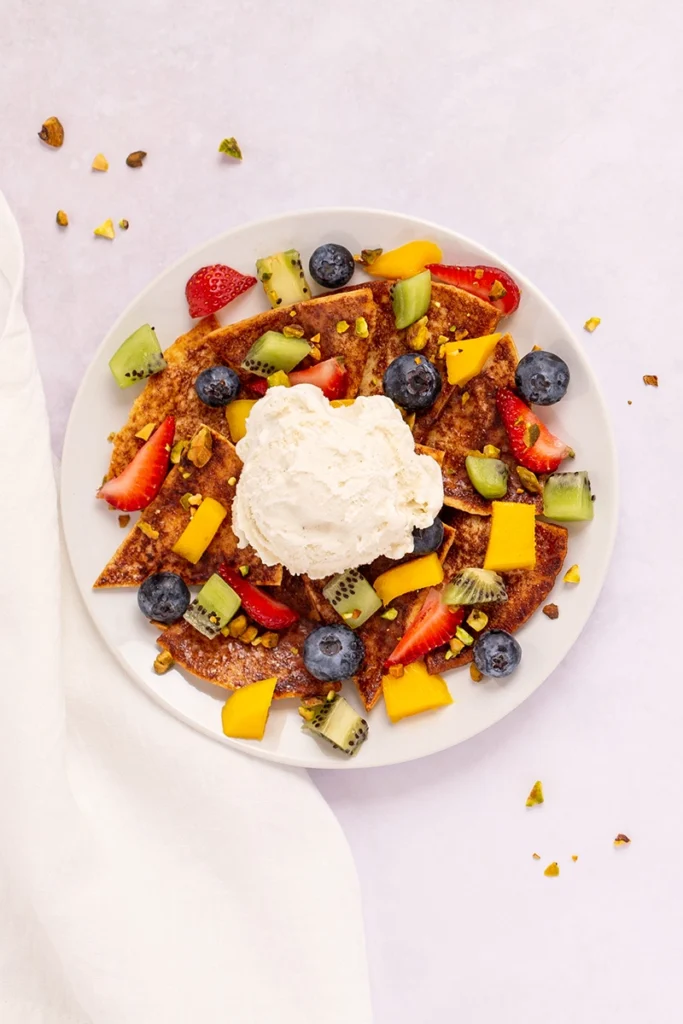 A plate of pancakes topped with mixed fruit pieces, nuts, and a scoop of vanilla ice cream offers a delightful twist reminiscent of fruit nachos, set on a light background with a white napkin. This tempting treat is perfect as fruit nachos for 1, beautifully combining breakfast comfort with fruity sweetness.