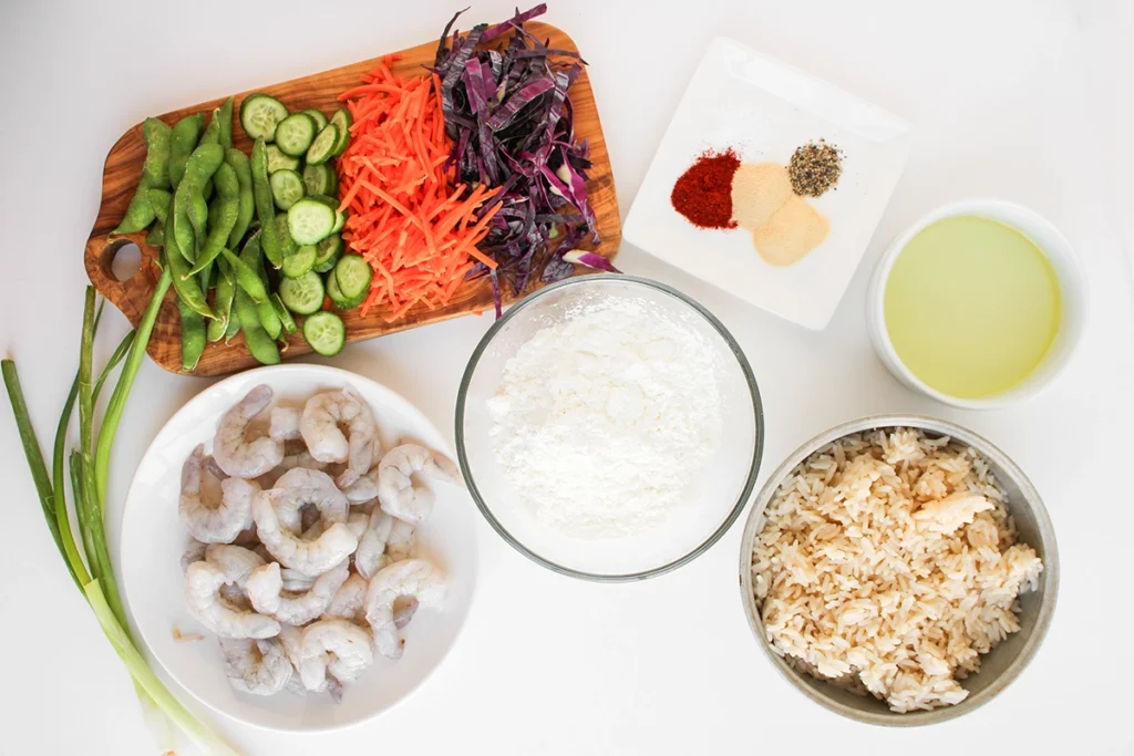 Raw shrimp, flour, cooked rice, sliced vegetables, powdered spices, oil, and green onions are arranged in separate bowls and plates on a white surface—perfect for preparing a flavorful bang bang shrimp bowl or trying out your favorite shrimp bowl recipe.