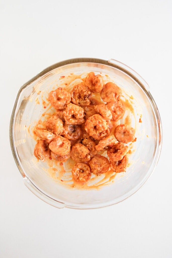 Glass bowl containing raw shrimp coated in a reddish marinade, placed on a white surface—perfect for preparing an easy shrimp bowl recipe or flavorful bang bang shrimp bowl.