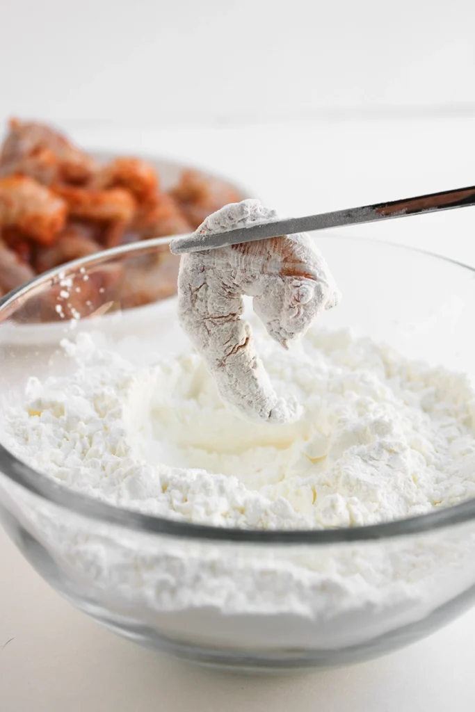 A shrimp is held with chopsticks over a bowl of flour or starch, coated and ready for frying in preparation for a delicious bang bang shrimp bowl. A plate of uncooked, seasoned shrimp is in the background, perfect for anyone searching for an easy shrimp bowl recipe.
