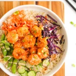 A flavorful bang bang shrimp bowl featuring rice, breaded shrimp tossed in a zesty orange sauce, sliced cucumber, snap peas, shredded carrots, and vibrant purple cabbage, all garnished with sesame seeds and green onions on a wooden board.