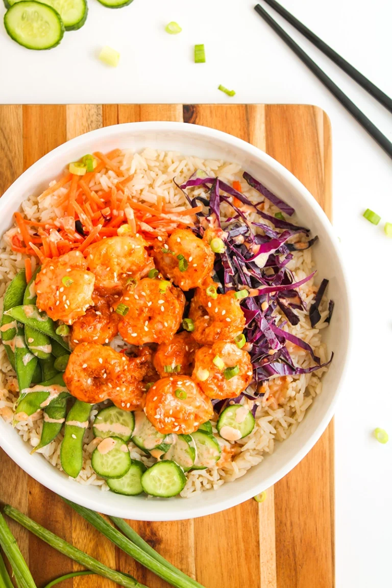 A flavorful bang bang shrimp bowl featuring rice, breaded shrimp tossed in a zesty orange sauce, sliced cucumber, snap peas, shredded carrots, and vibrant purple cabbage, all garnished with sesame seeds and green onions on a wooden board.
