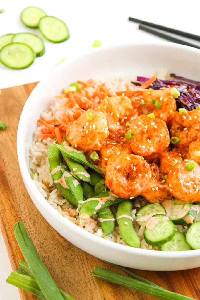 A white bowl featuring a colorful bang bang shrimp bowl: tender shrimp in a flavorful sauce, served over rice and surrounded by snap peas, shredded carrots, purple cabbage, and sliced cucumbers. Finished with a drizzle of creamy sauce and garnished with sesame seeds and green onions, this shrimp bowl recipe is as vibrant as it is delicious.