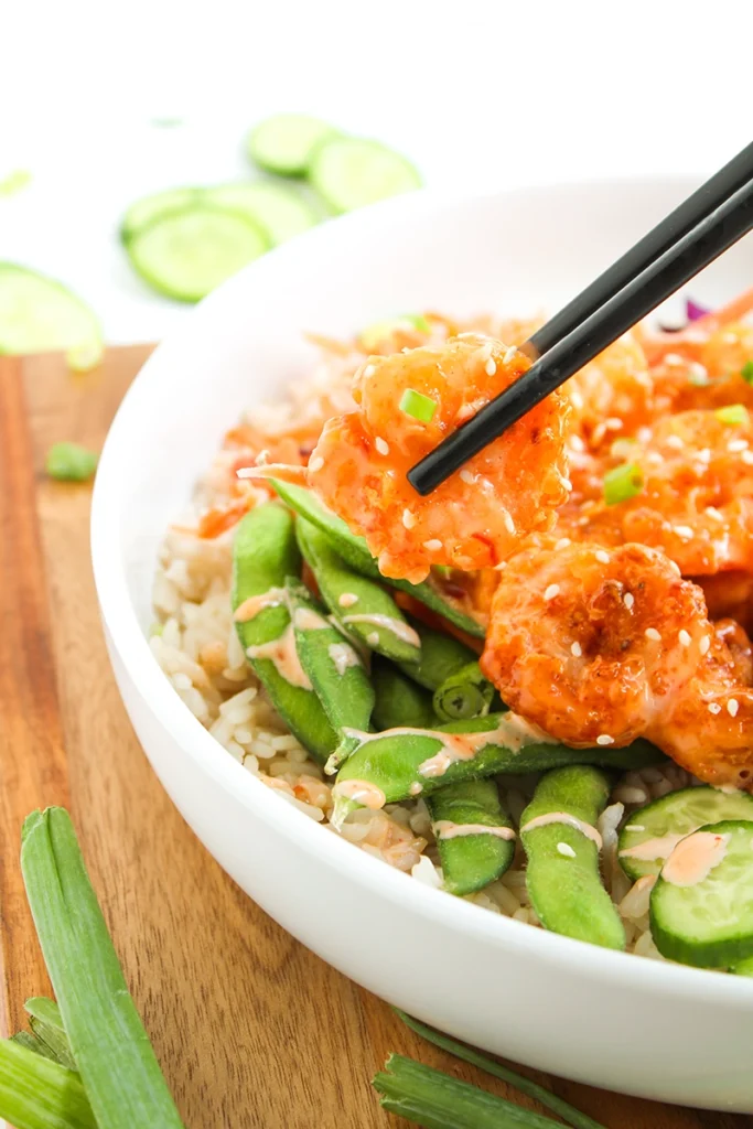 A bowl of rice topped with breaded shrimp, snap peas, and sauce, with chopsticks picking up a shrimp creates the perfect bang bang shrimp bowl. Sliced cucumbers and green onions are on the side, making this shrimp bowl recipe both delicious and vibrant.