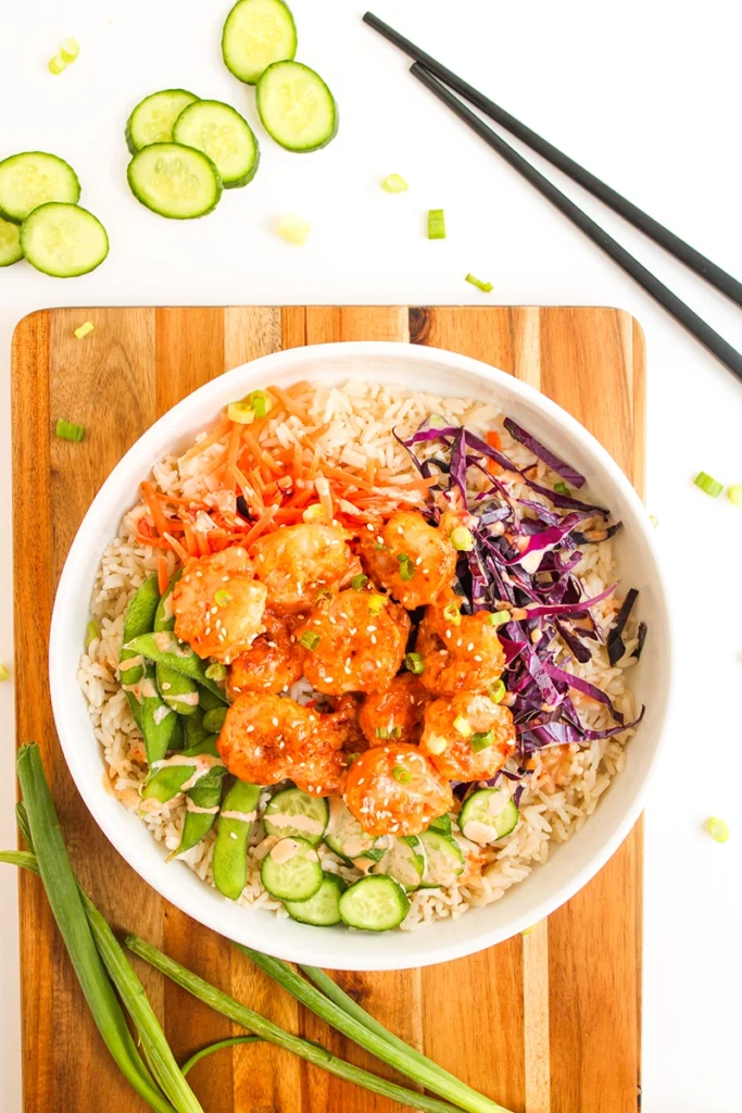 A delicious shrimp bowl recipe featuring a bowl of rice topped with sliced cucumber, avocado, red cabbage, carrots, and sauced shrimp, served on a wooden board with chopsticks and green onions nearby.