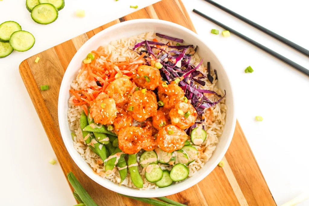 A bang bang shrimp bowl featuring a bowl of rice topped with shrimp in sauce, sliced cucumber, snap peas, shredded red cabbage, and julienned carrots, served on a wooden board with chopsticks. This flavorful shrimp bowl recipe makes for a colorful and delicious meal.