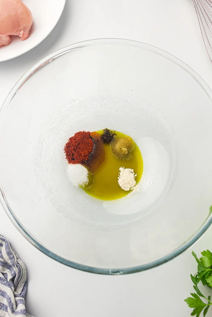 A glass bowl with olive oil, paprika, salt, pepper, onion powder, and other spices on a white surface, ready for mixing—perfect for seasoning your air fryer chicken breast before cooking.