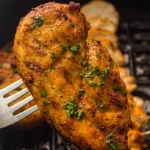 A cooked, seasoned air fryer chicken breast garnished with chopped herbs is held by a fork over an air fryer basket with more pieces of air fried chicken in the background.