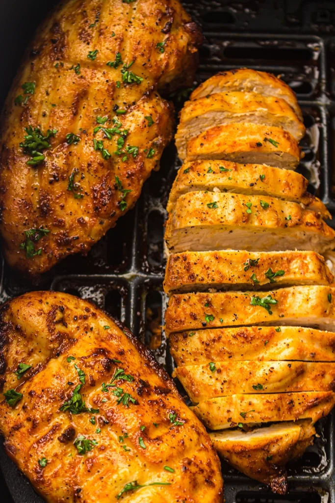 Three seasoned, cooked air fryer chicken breasts, one sliced, garnished with chopped herbs, placed in a black grill pan.