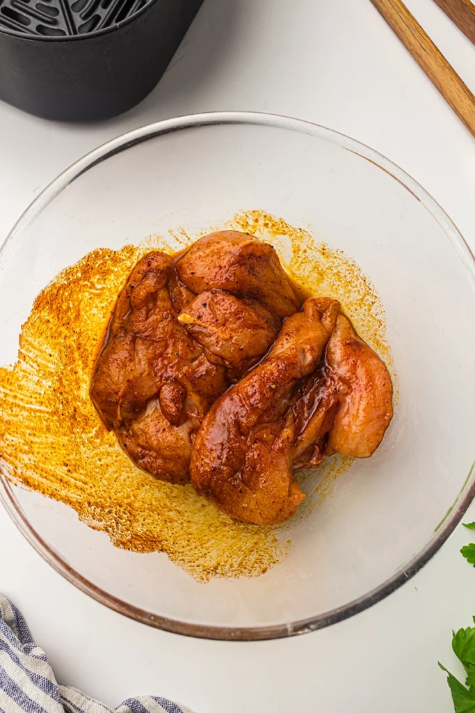 Raw chicken breasts coated in a reddish marinade sit in a glass bowl on a white surface, ready to be transformed into flavorful air fryer chicken breast, with seasoning spread along the sides of the bowl.