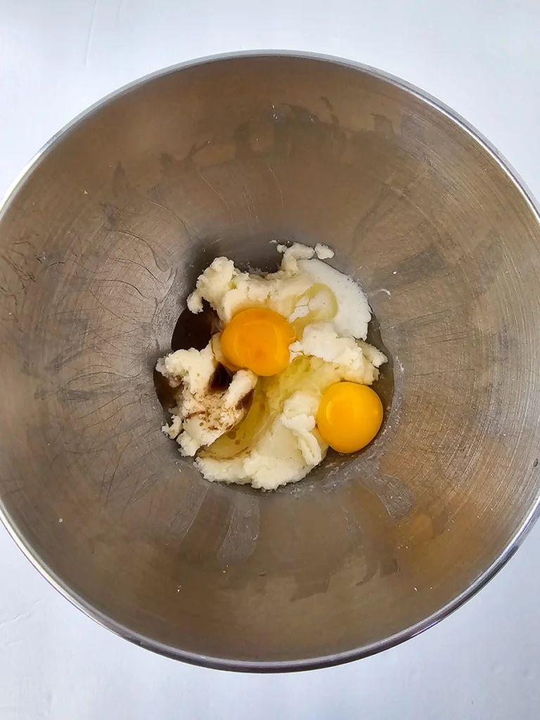 A stainless steel mixing bowl containing two raw eggs, sugar, vanilla extract, softened butter, and ripe bananas sits ready to be mixed for a delicious small batch banana bread.