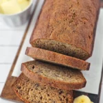 A loaf of small batch banana bread partially sliced on a wooden board, with a ramekin of butter and banana slices beside it.