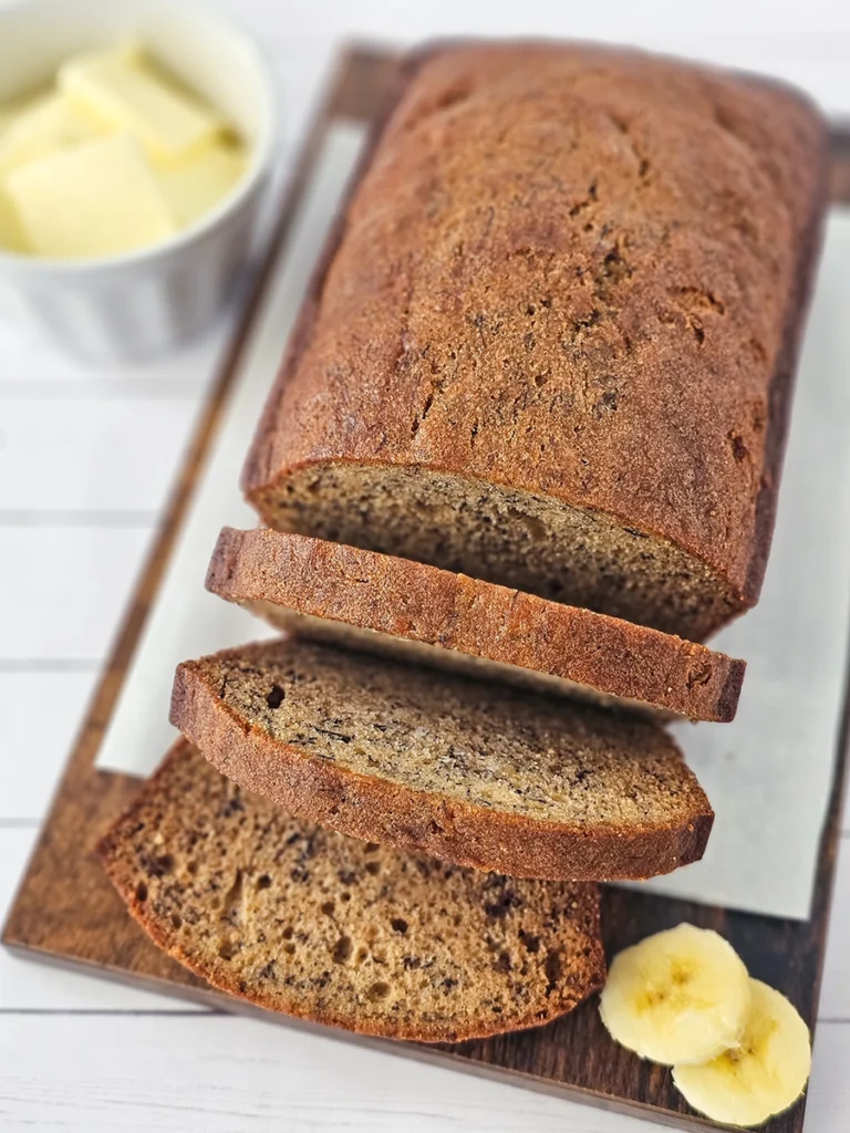 A loaf of small batch banana bread partially sliced on a wooden board, with a ramekin of butter and banana slices beside it.