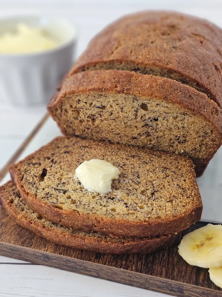 A loaf of small batch banana bread with two slices cut, one topped with a dollop of butter. Slices rest on a wooden board with banana pieces nearby. A dish of butter is in the background.