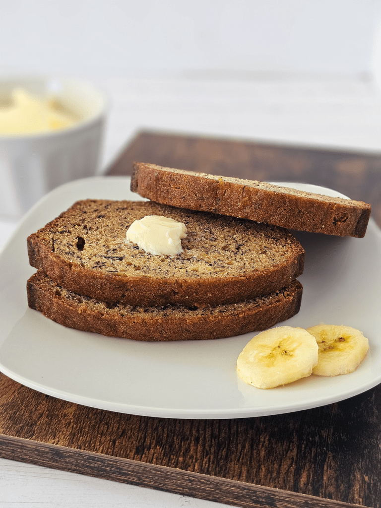 Two slices of small batch banana bread with butter on a white plate, accompanied by two banana slices; a bowl of butter is in the background.