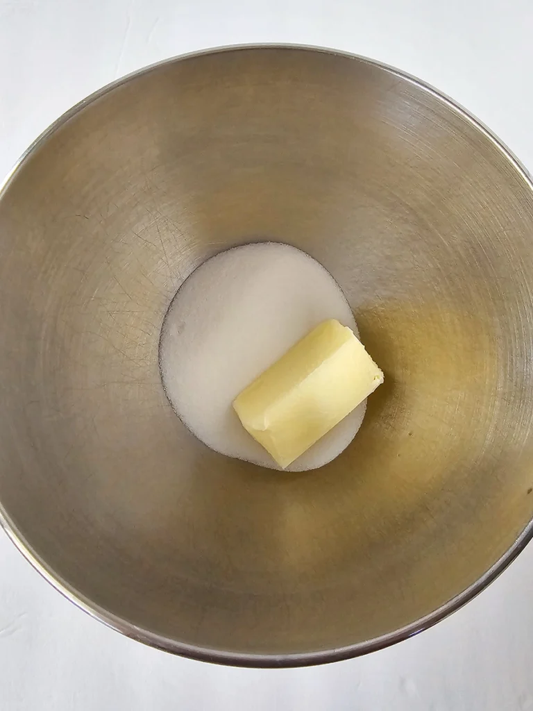 A metal mixing bowl containing a stick of butter and a pile of granulated sugar on a white background, ready to be used for making small batch banana bread.