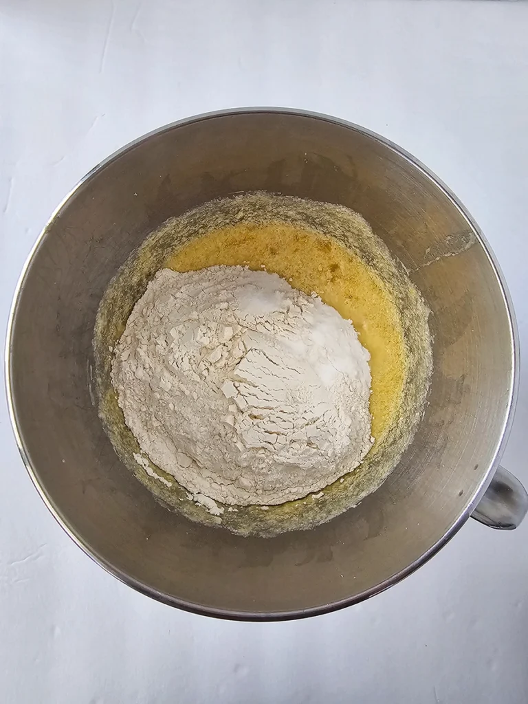 A metal mixing bowl containing flour on top of a yellow liquid mixture for small batch banana bread, placed on a white surface, viewed from above.