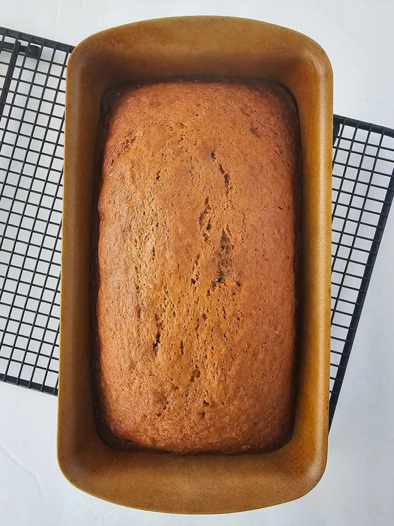 A loaf of small batch banana bread sits in a brown loaf pan on top of a black cooling rack.